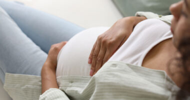 A pregnant woman sits on a sofa while cradling her belly with both hands.