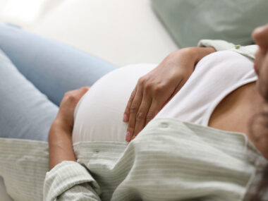 A pregnant woman sits on a sofa while cradling her belly with both hands.