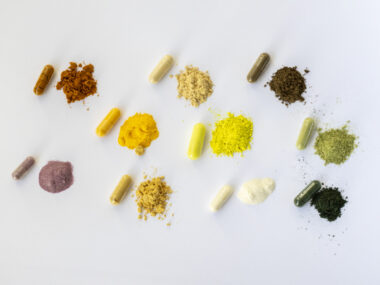 Assorted colorful capsules and small piles of powdered supplements arranged in rows on a white background.