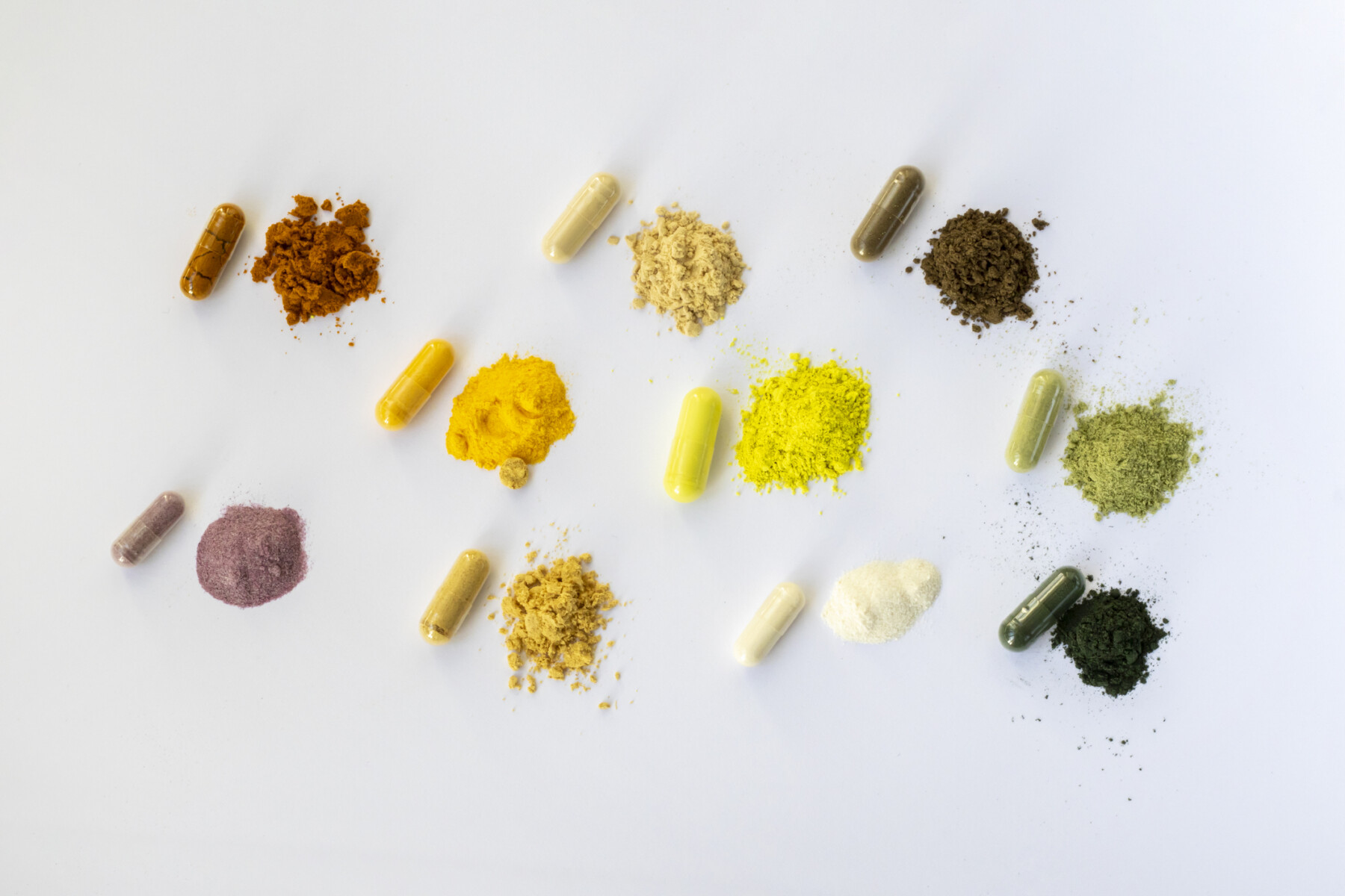 Assorted colorful capsules and small piles of powdered supplements arranged in rows on a white background.