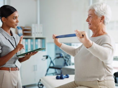 Healthcare professional holding a tablet guides an older woman performing resistance band exercises in a rehabilitation setting.