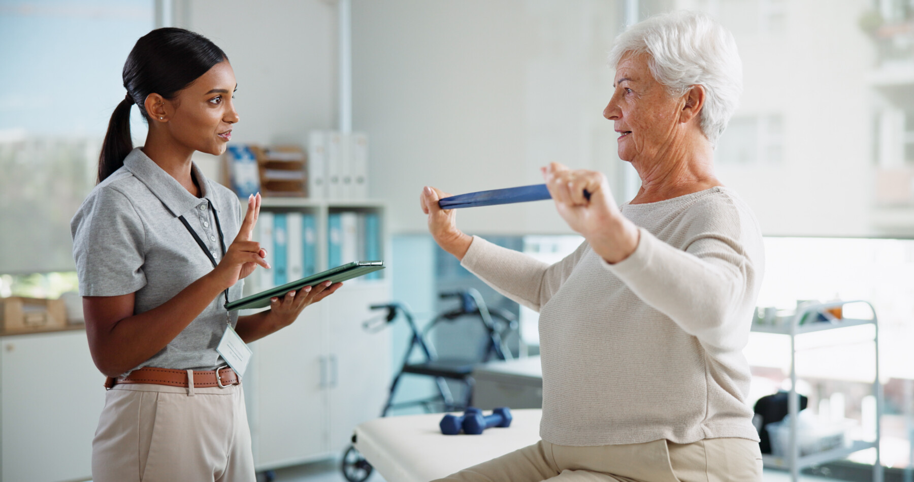 Healthcare professional holding a tablet guides an older woman performing resistance band exercises in a rehabilitation setting.