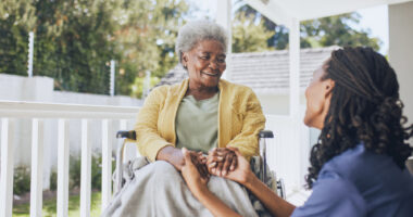 A young female nurse holds the hands of an older patient in a wheelchair.