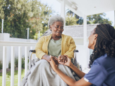 A young female nurse holds the hands of an older patient in a wheelchair.