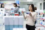 A woman holds a medication package in each hand, comparing them, while visiting a pharmacy.