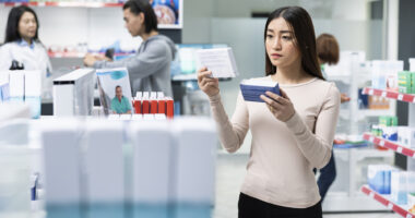A woman holds a medication package in each hand, comparing them, while visiting a pharmacy.