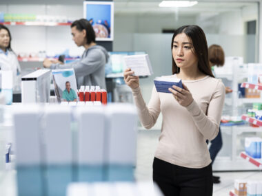 A woman holds a medication package in each hand, comparing them, while visiting a pharmacy.