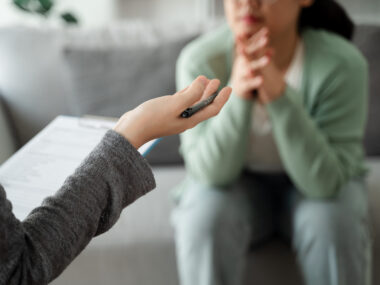 A patient sits on a couch listening to a psychologist holding a clipboard.