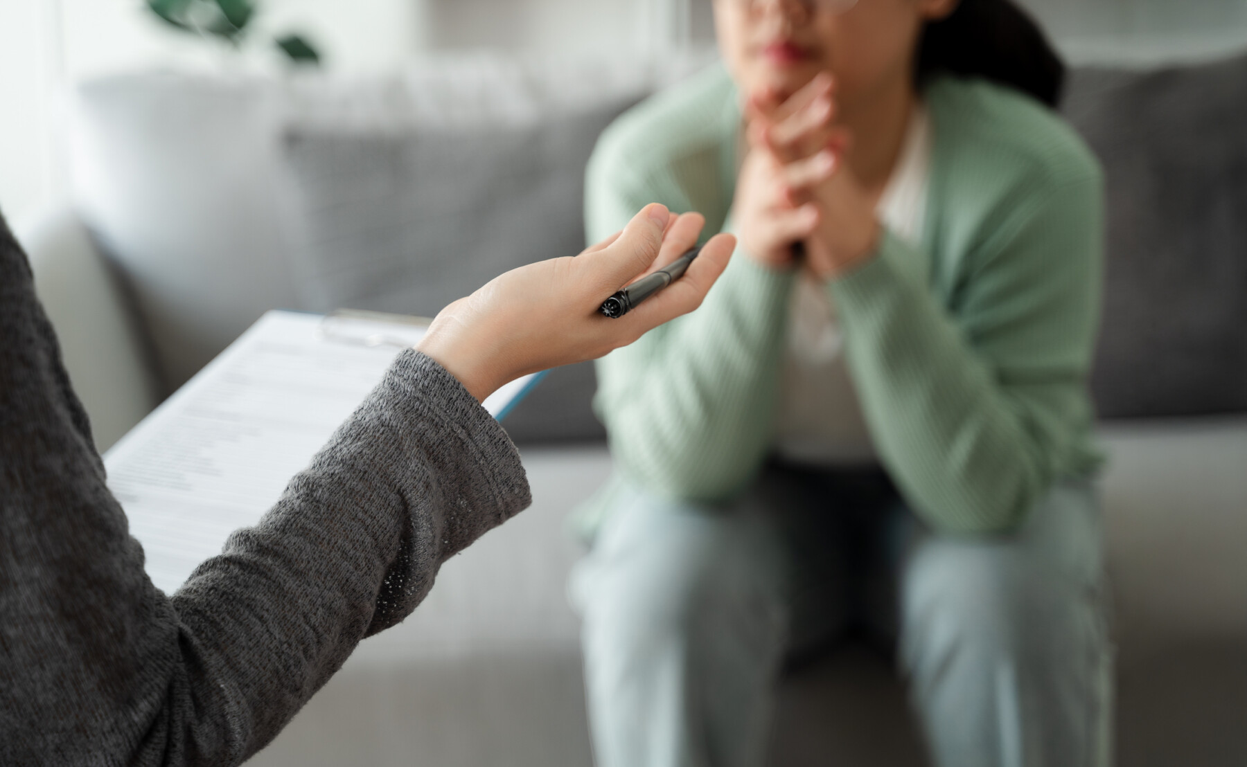 A patient sits on a couch listening to a psychologist holding a clipboard.